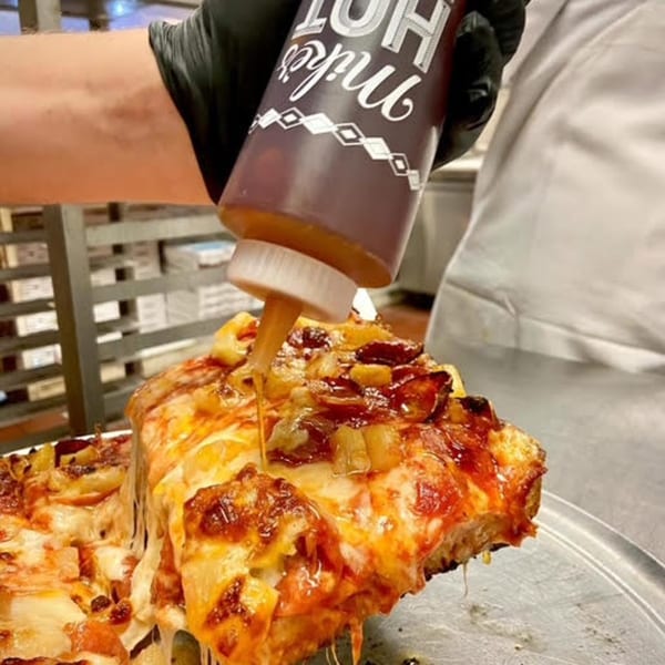 A hand in a black glove squeezes hot honey onto a cheesy slice of Franco's Italian pizza.
